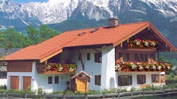 Una cabaña tradicional con techo inclinado, balcones de madera con jardineras y montañas al fondo.