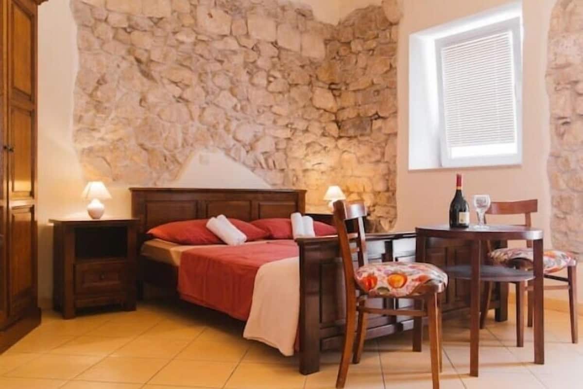 A bedroom with a stone wall, a bed with red bedding, a wooden nightstand with a lamp, a wooden chair, and a table with a bottle and glass.