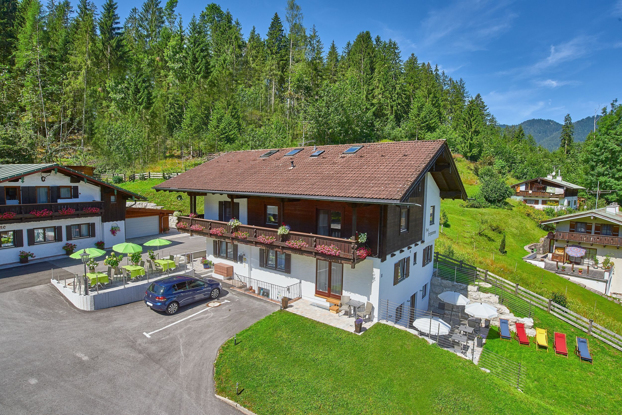 A two-story chalet with a balcony, a parking area, and a small outdoor seating area with umbrellas.
