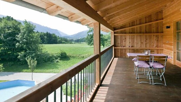 A wooden balcony with a view of a green field and mountains. There are two tables with chairs.
