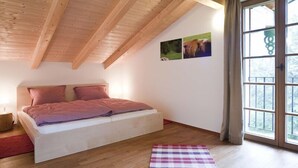 A bedroom with a wooden ceiling, a bed with pink bedding, a checkered rug, and a window with curtains.