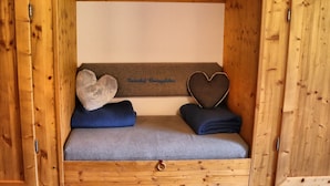 A wooden cabin interior with a bed, two heart-shaped pillows, and a bench with a name plaque.