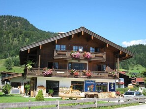 Eine Holzhütte mit Balkonen, die mit Blumen geschmückt sind, ein Geschäft mit der Aufschrift "hausberg" und eine Aussicht auf einen grünen Berg.