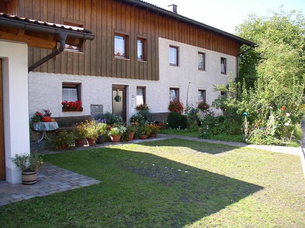 A two-story building with a wooden facade, multiple windows, and a well-maintained garden with various potted plants and flowers. - Ferienwohnung 2 im Erdgeschoss, 65 qm, für 2 bis 4 Personen, 2 sep. Schlafzimmer, bedingt geeignet für Rollstuhlfahrer (Grabenstätt)
