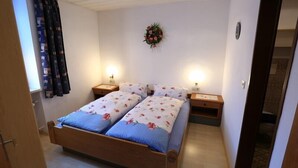A bedroom with a wooden bed, two bedside tables with lamps, a floral wall decoration, and a window with curtains. - Zwei-Raum-Ferienwohnung (1) "Parterre" 28qm, Dusche/WC, Extra-Schlafraum, Küche (Reit im Winkl)