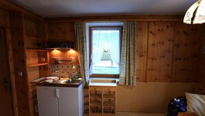 A compact kitchen with wooden walls, a built-in stove, a sink, and a small window with curtains. - Zwei-Raum-Ferienwohnung (1) "Parterre" 28qm, Dusche/WC, Extra-Schlafraum, Küche (Reit im Winkl)
