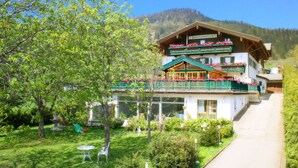 Ein traditionelles Gebäude im alpinen Stil mit einem Balkon, der mit Blumen geschmückt ist, umgeben von üppiger Vegetation und einer Bergkulisse.