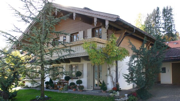 A two-story wooden house with a balcony, surrounded by potted plants and trees. - Ferienwohnung, 60 qm, 1 Schlafzimmer, Balkon (Siegsdorf)