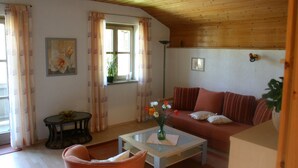 A cozy living room with a wooden ceiling, a sofa, a coffee table with a vase of flowers, a small table with a plant, and a window with curtains. - Ferienwohnung, 60 qm, 1 Schlafzimmer, Balkon (Siegsdorf)