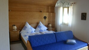 A bedroom with a wooden ceiling, a bed with blue bedding, a nightstand with a lamp, and a window with curtains. - Ferienwohnung, 60 qm, 1 Schlafzimmer, Balkon (Siegsdorf)