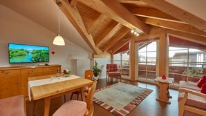 Ein großzügiges Loft mit hohen Decken, Holzbalken, einer Essecke mit Tisch und Stühlen, einem Fernseher und großen Fenstern mit Blick ins Freie.