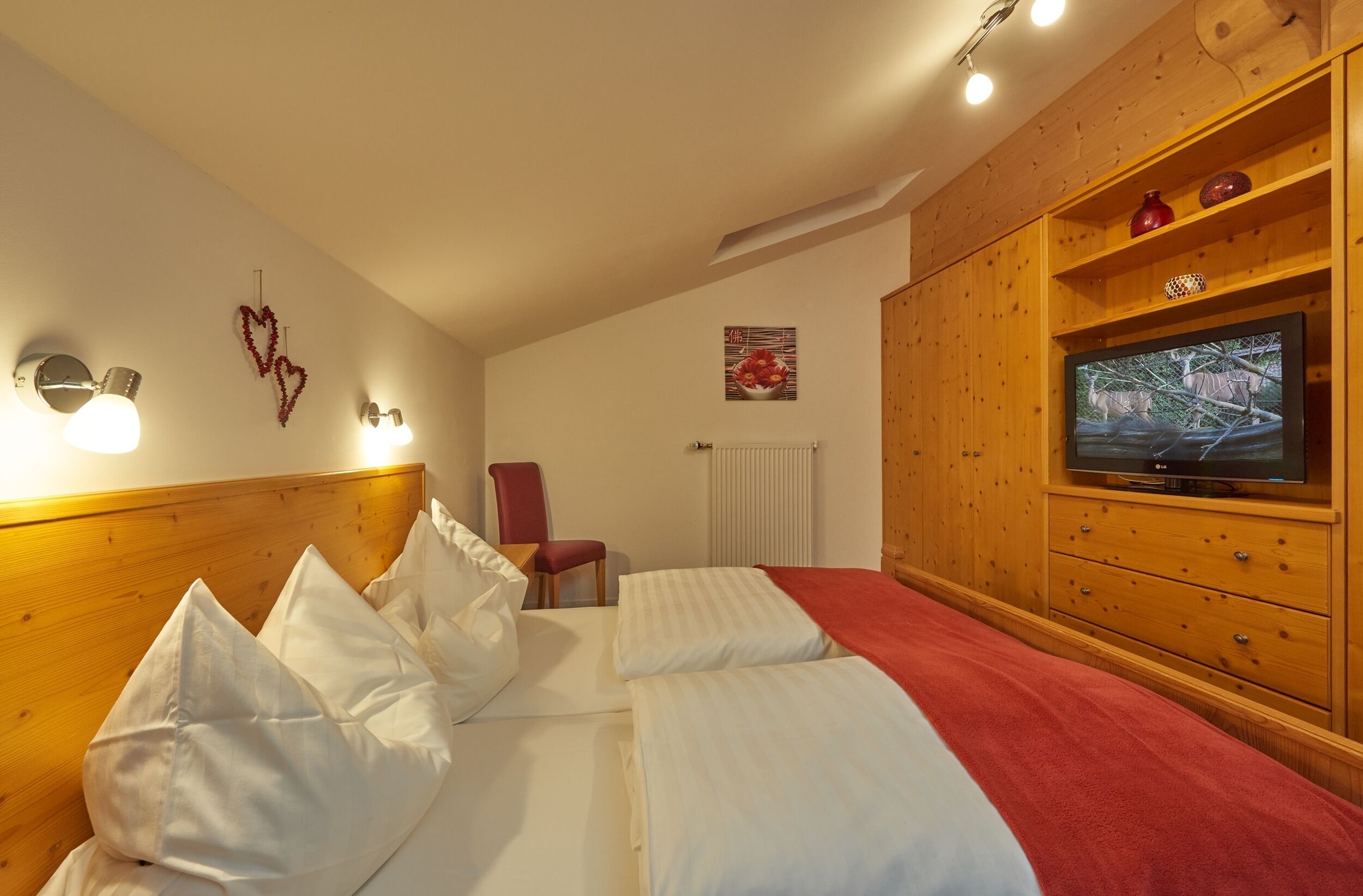 A hotel room with a wooden headboard, a bed with white linens, a red chair, a television on a wooden cabinet, and wall-mounted lights.
