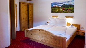 Un lit en bois avec du linge de lit blanc, une armoire en bois et un tableau représentant des vaches sur une colline.