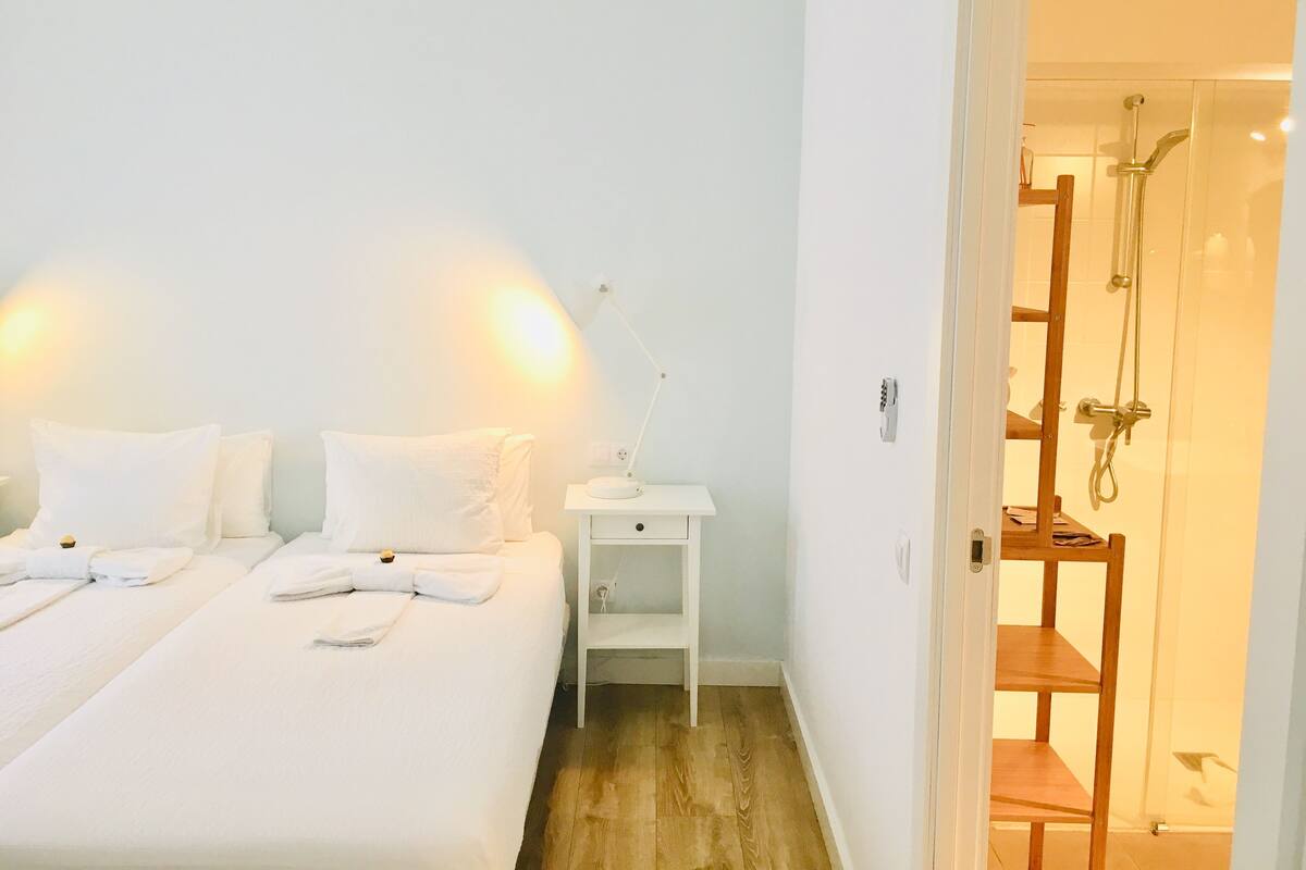 Ein Hotelzimmer mit zwei Betten, einem Nachttisch mit Lampe, einem Holzboden und einem sichtbaren Duschbereich durch eine offene Tür.
