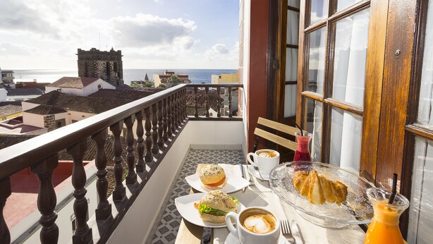 Un balcón con una mesa dispuesta para el desayuno, que incluye un sándwich, café y jugo.