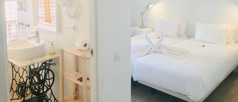 Habitación de hotel con baño que incluye un lavabo, espejo y toallero. El dormitorio tiene una cama, mesitas de noche y una lámpara colgante.