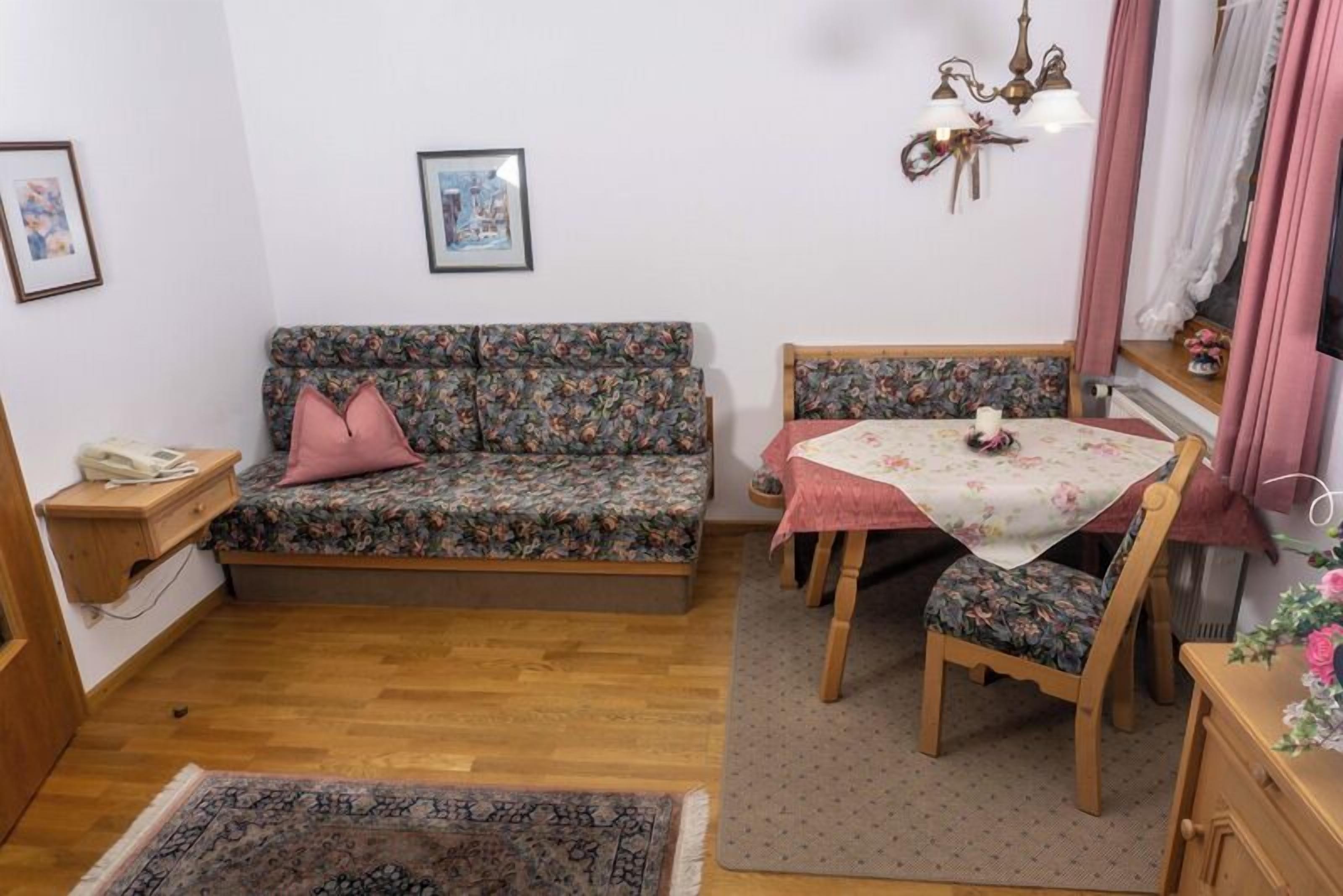 Una habitación acogedora con un sofá floral, una mesa de comedor con sillas, una alfombra pequeña y un teléfono sobre una mesita de madera.