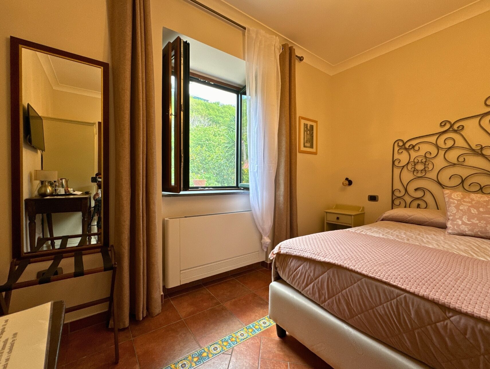 A bedroom with a bed, a bedside table, a mirror, a window with curtains, and a radiator.