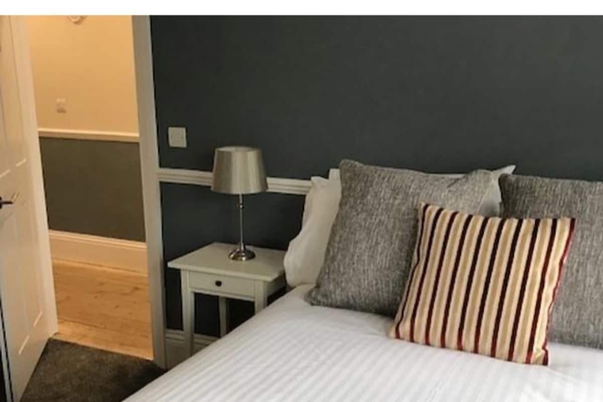 A bedroom with a bed, two pillows, a bedside table with a lamp, and a door leading to another room.