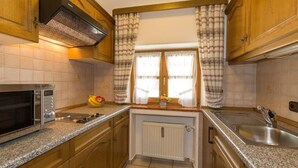 Eine kleine Küche mit Holzschränken, einer eingebauten Mikrowelle, einer Spüle und einem Fenster mit gemusterten Vorhängen.
