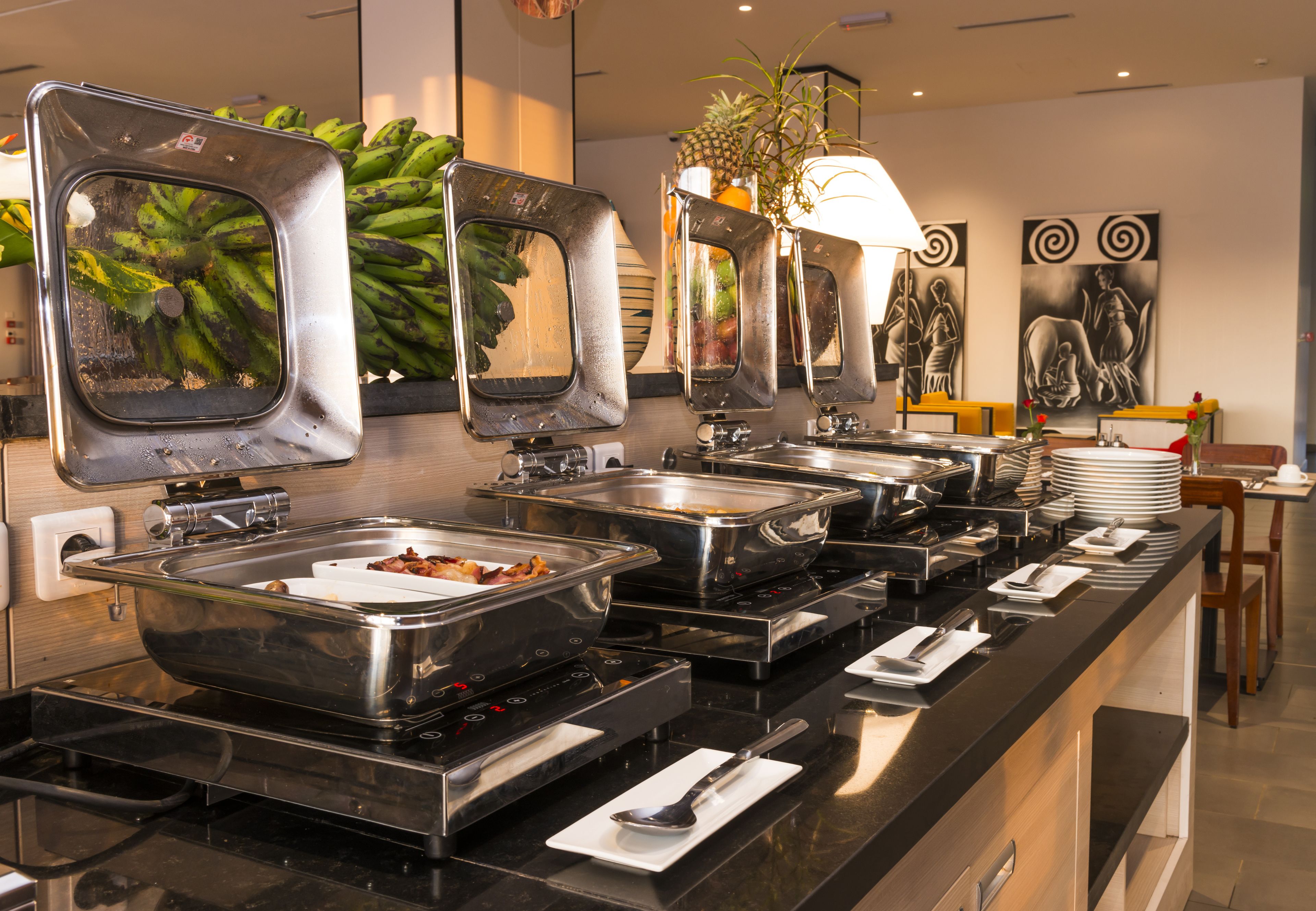 A buffet setup with multiple serving stations, a potted plant, and artwork on the wall.