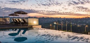 An infinity pool with lounge chairs and umbrellas, overlooking a cityscape at sunset. - ONOMO Hotel Kigali (Kigali)