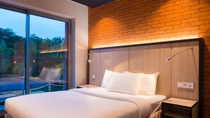 Une chambre moderne avec un grand lit, une table de chevet, une lampe fixée au mur et une porte en verre donnant sur un espace extérieur.