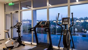 Une salle de sport équipée de machines de cardio, d’un banc de musculation et offrant une vue sur le paysage urbain grâce à de grandes fenêtres.