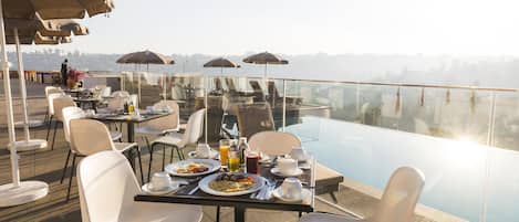 A rooftop dining area with tables, chairs, and umbrellas overlooking a pool.
