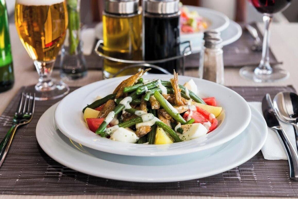 A plate of food with green beans, tomatoes, and a white sauce, accompanied by a glass of beer and salt and pepper shakers.