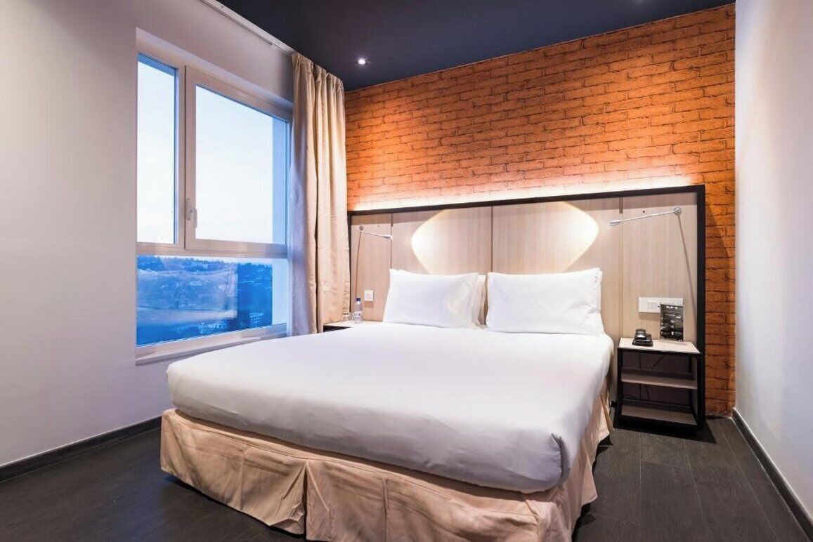 Ein Hotelzimmer mit einem großen Bett, einem Fenster mit Vorhängen, einer Ziegelfassade und einem Nachttisch mit Lampe.