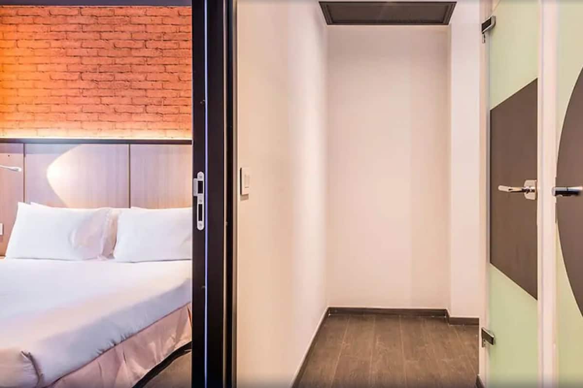 Camera d'albergo moderna con parete in mattoni, un letto grande con lenzuola bianche, un corridoio stretto e una porta rotonda.