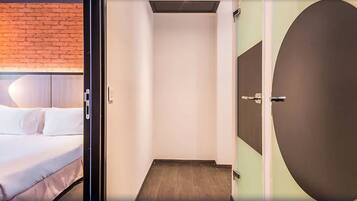 Una habitación de hotel moderna con una pared de acento de ladrillo, una cama grande con ropa blanca y un estrecho pasillo con una puerta redonda.