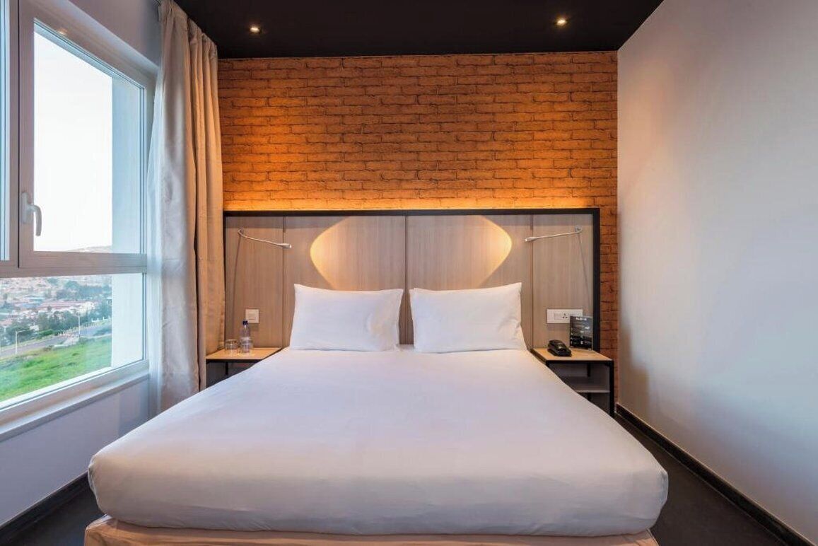 A hotel room with a brick wall, a large bed, two bedside tables, and a window with curtains.