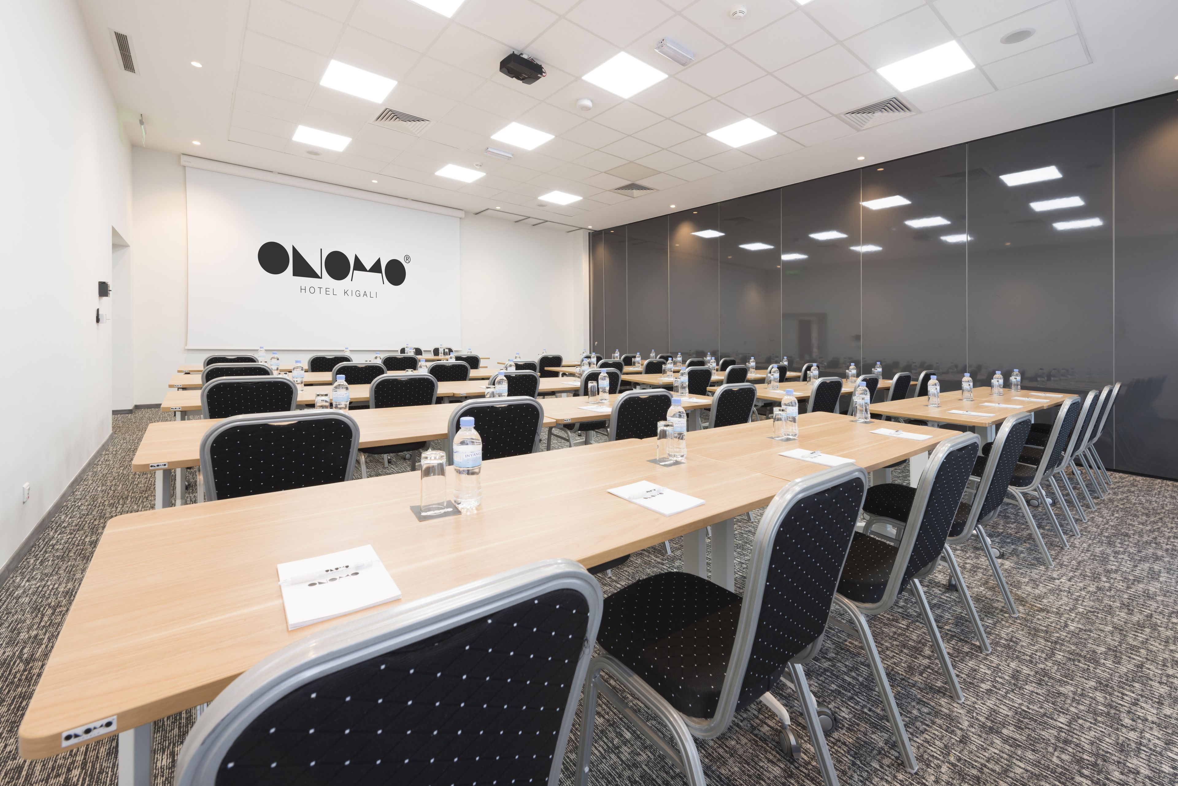 A conference room with multiple tables and chairs, equipped with water bottles and papers, and a large screen on the wall.