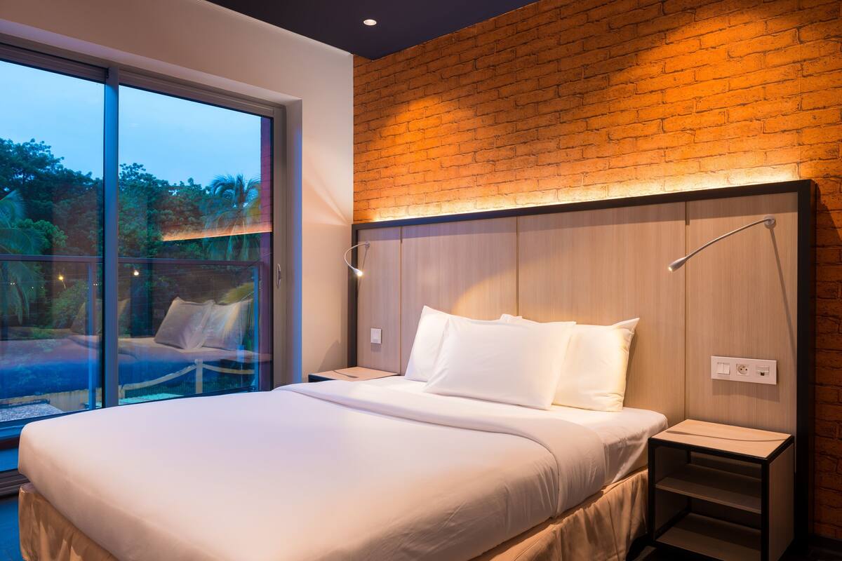 Una camera da letto moderna con un letto grande, un comodino, una lampada a parete e una porta scorrevole in vetro che dà su uno spazio esterno.