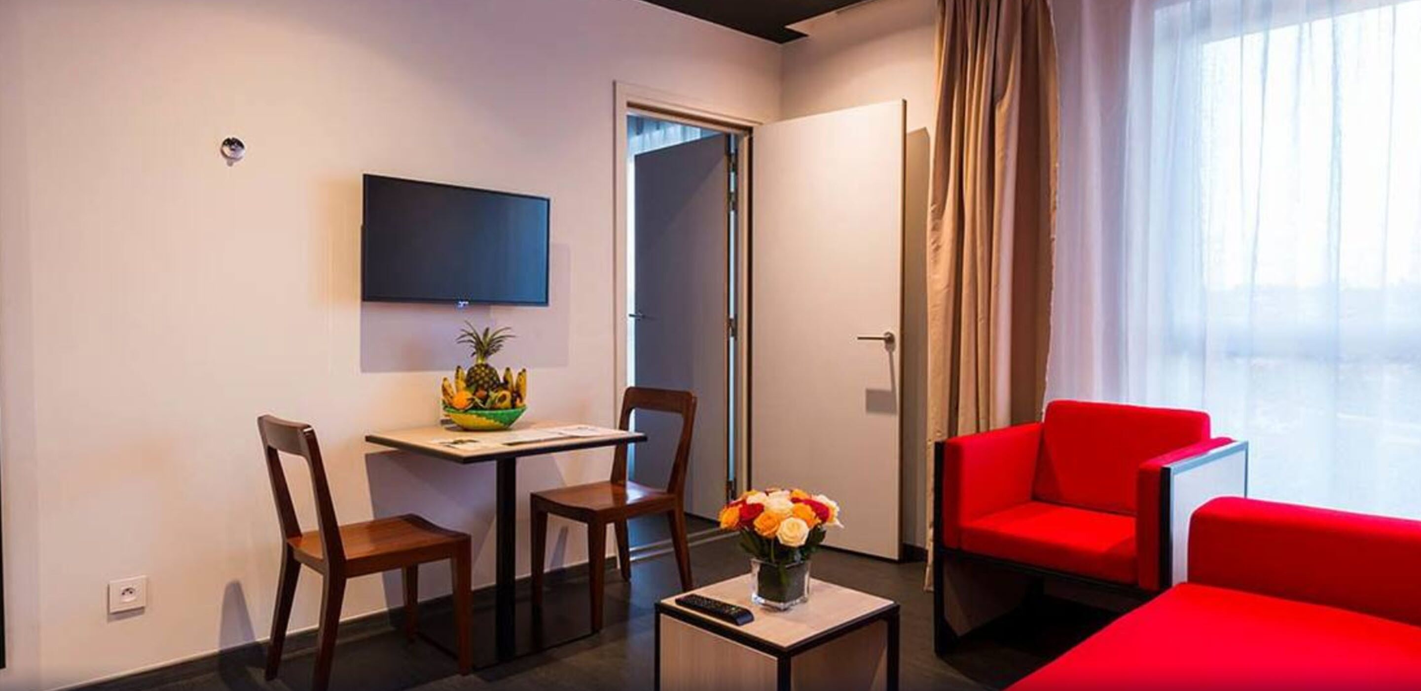 Ein Hotelzimmer mit Fernseher, einem Esstisch mit Stühlen, einem roten Sofa und einem Fenster mit Vorhängen.