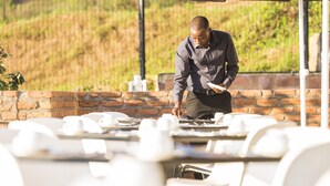 Un homme, vêtu d’une chemise et d’un pantalon sombres, se tient près d’une table sur laquelle est posé un ordinateur portable. Il surplombe une salle à manger avec des chaises et des tables blanches, le tout adossé à un mur de briques et à une colline verdoyante.