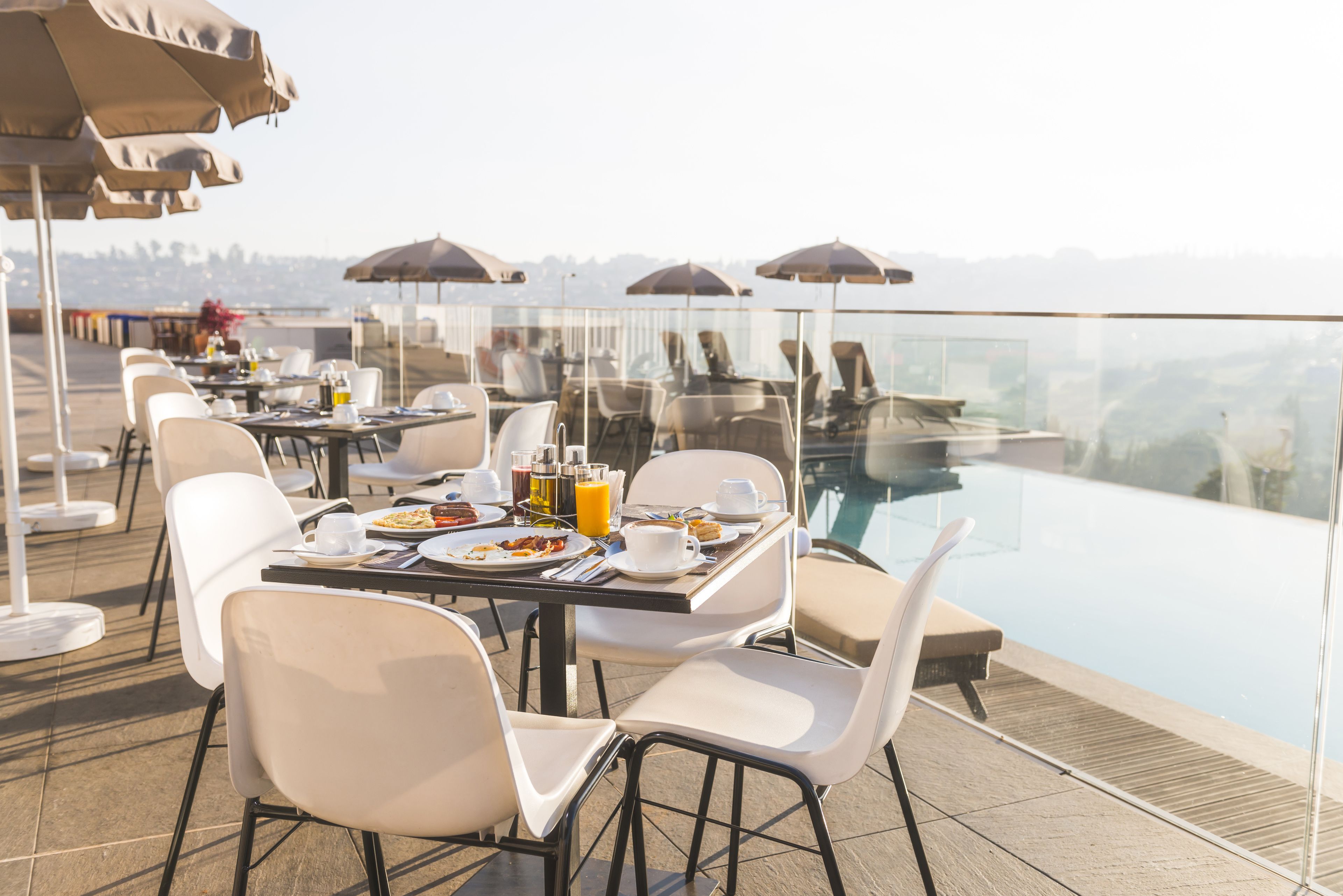A rooftop dining area with tables, chairs, and umbrellas overlooking a pool.