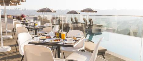 A rooftop dining area with tables, chairs, and umbrellas overlooking a pool.