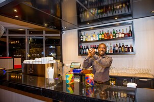 Una zona de bar con estantes llenos de botellas de licor, una máquina de café y una barra con tazas y un vaso de colores.