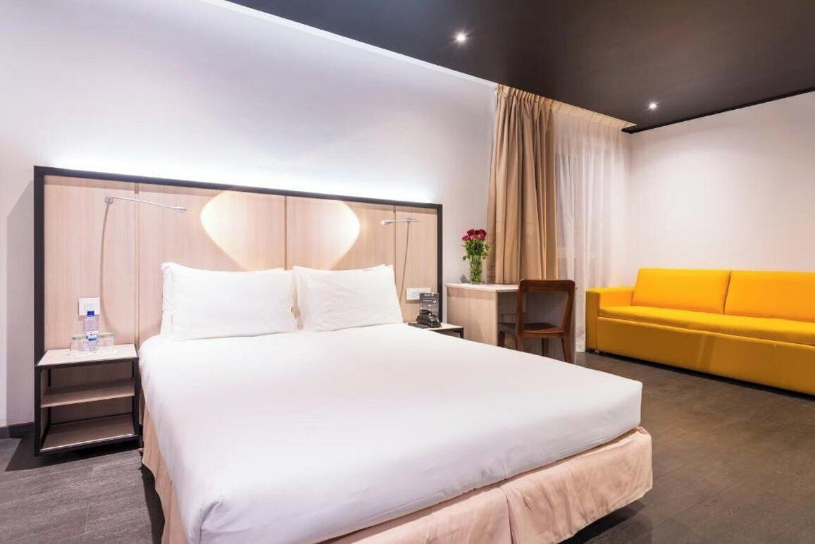 A hotel room with a large bed, a desk, a chair, a yellow sofa, and a window with curtains.