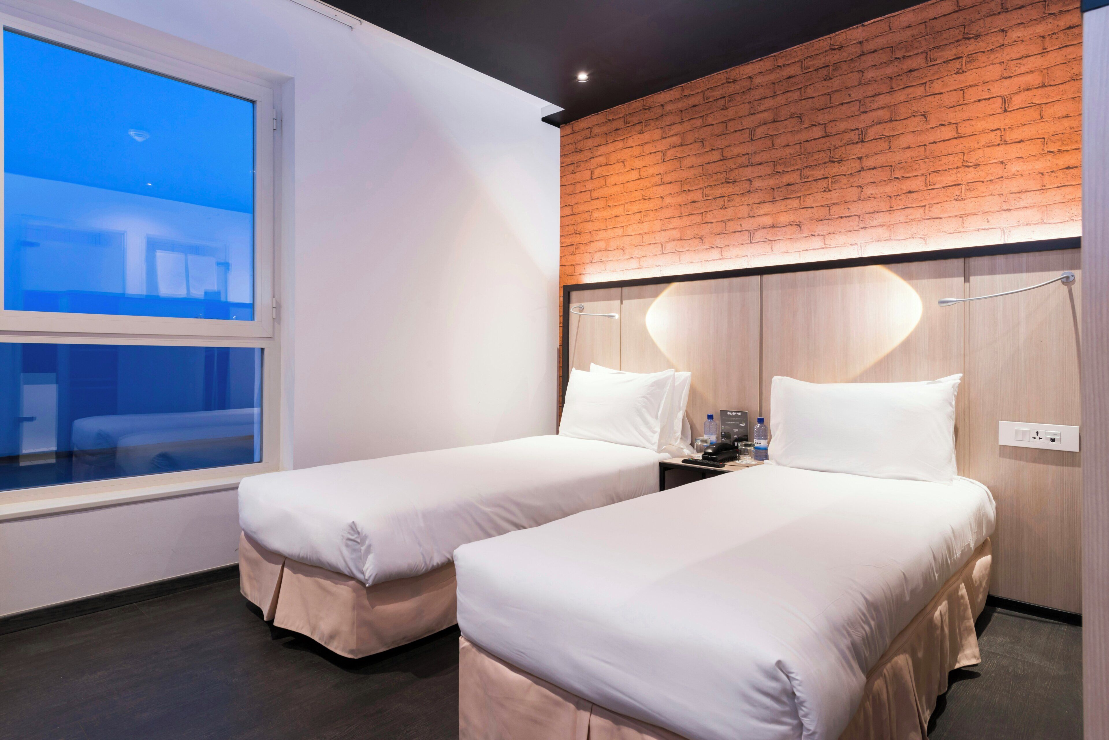 A hotel room with two single beds, a large window, a brick accent wall, and a wooden headboard.