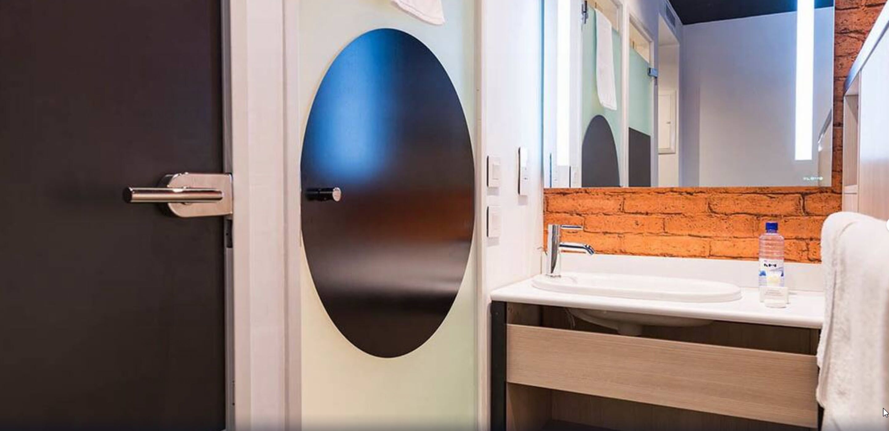 Un baño moderno con puerta oscura, espejo grande, lavamanos blanco y pared de ladrillo decorativa.