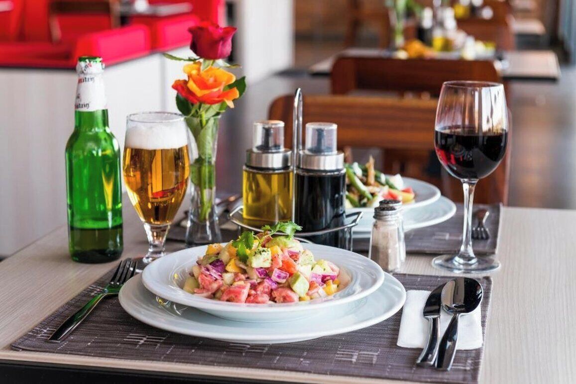 A dining table with a plate of food, a glass of beer, a bottle of beer, a glass of red wine, and condiments.
