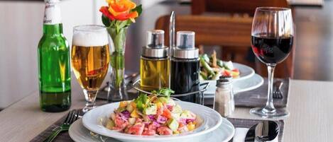 A dining table with a plate of food, a glass of beer, a bottle of beer, a glass of red wine, and condiments.