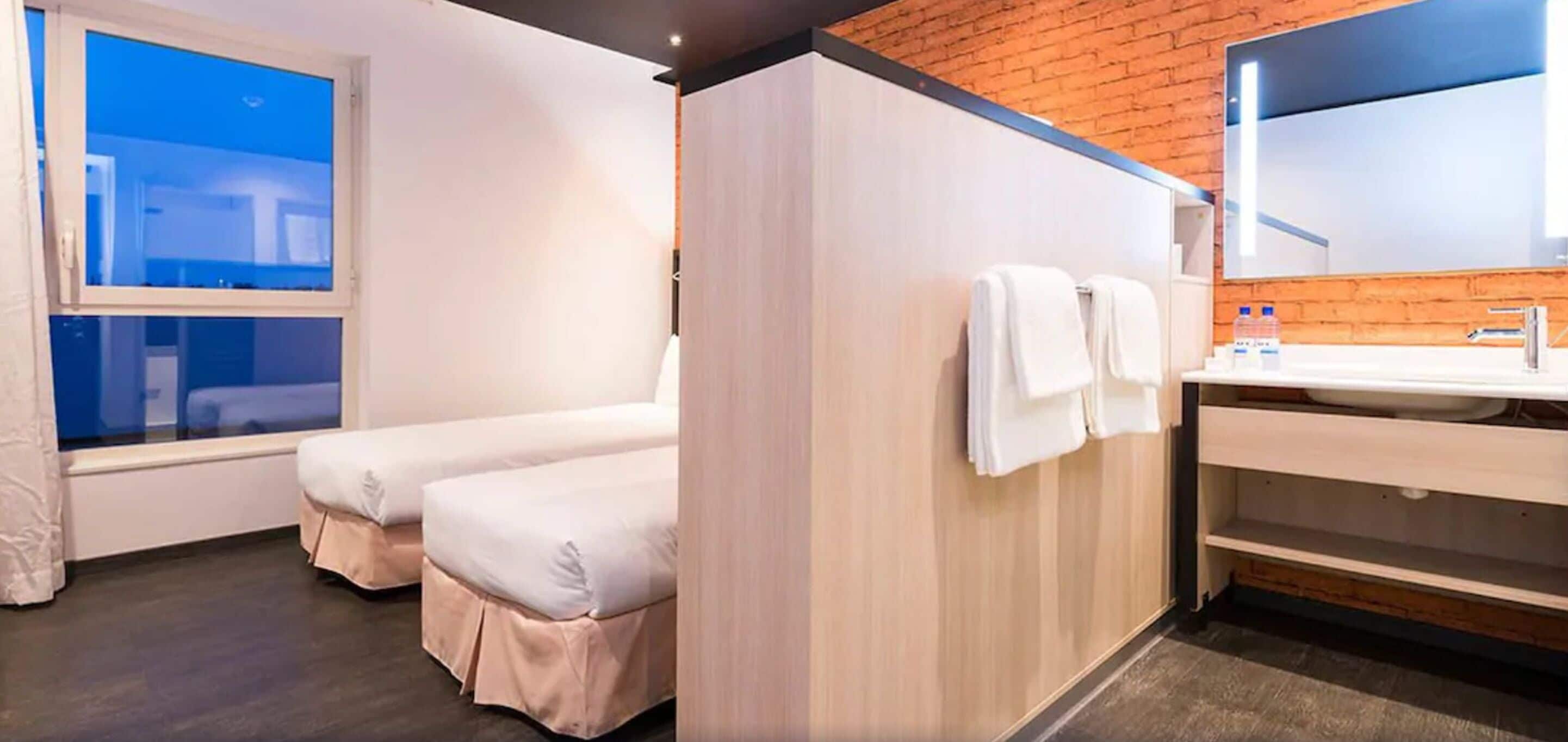 A hotel room with two beds, a large mirror, a sink, and a window with a view.