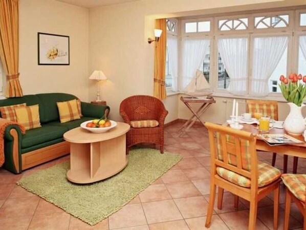 Una sala de estar con un sofá verde, una mesa redonda de centro, un sillón de mimbre, una mesa de comedor con sillas y una ventana con cortinas.