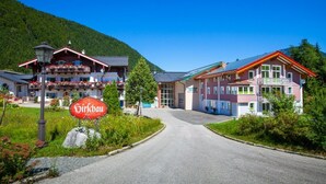A hotel with a red sign, a mountain backdrop, and a clear sky. - Kehlstein, 1-2 Pers., 80 m², Schlafzimmer, Wohnzimmer, Kochnische, Badewanne, Dusche und WC, Terrasse (Bischofswiesen)