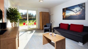 A modern living room with a wooden coffee table, a grey sofa with red cushions, a TV on a wooden stand, and a large window with a view of a garden and mountains. - Kehlstein, 1-2 Pers., 80 m², Schlafzimmer, Wohnzimmer, Kochnische, Badewanne, Dusche und WC, Terrasse (Bischofswiesen)